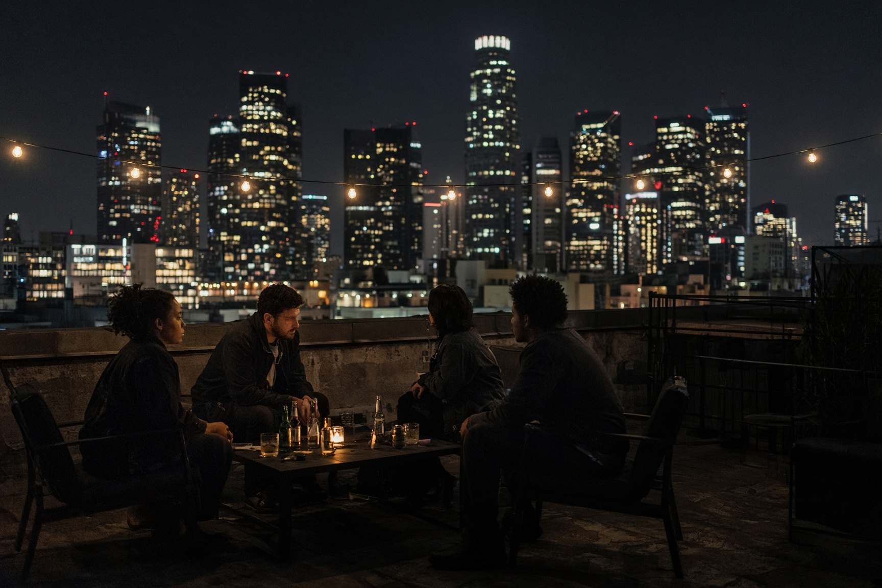 Four people seated around a low table on a rooftop at night, city towers behind them
